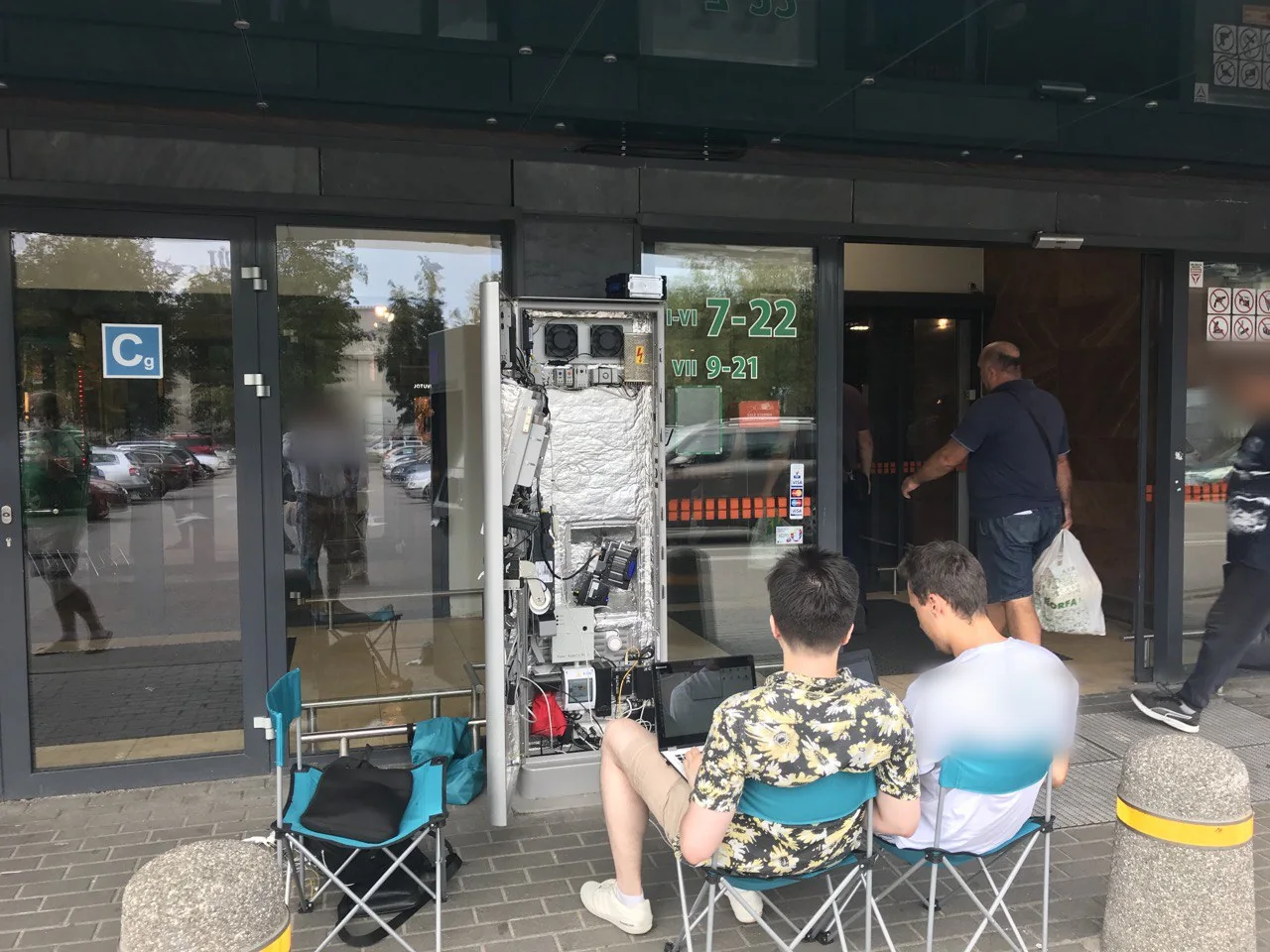 Terminal parkingowy przed supermarketem w UE podczas uruchomienia: płatności, sterowanie i łączność w otwartej obudowie. Przechodnie i nadruki są rozmyte.